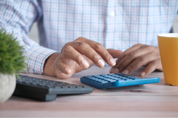 Person's hand about to use a blue calculator that is on a wood desk along with a computer keyboard, potted plant, and yellow mug.