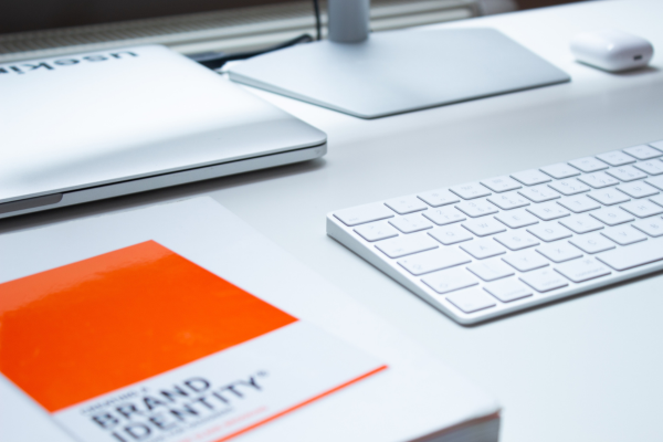 Table with desktop computer, wireless keyboard and orange notebook with brand identity  written on it.