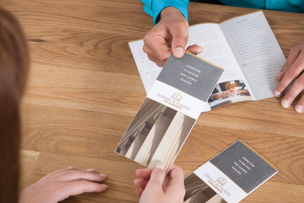 Person handing brochure to someone else over a wood desk